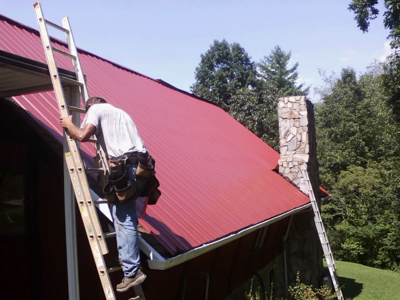 Roofer on a ladder beside a red metal roof and stone chimney in Grand Meadow